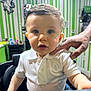 toddler, child, boy, white_polo_shirt, haircut, barber_chair, adult_hand, blue_eyes, short_hair, indoor, green_wall, striped_wall, mirror, salon, person, portrait, curious, candid, clothing, furniture