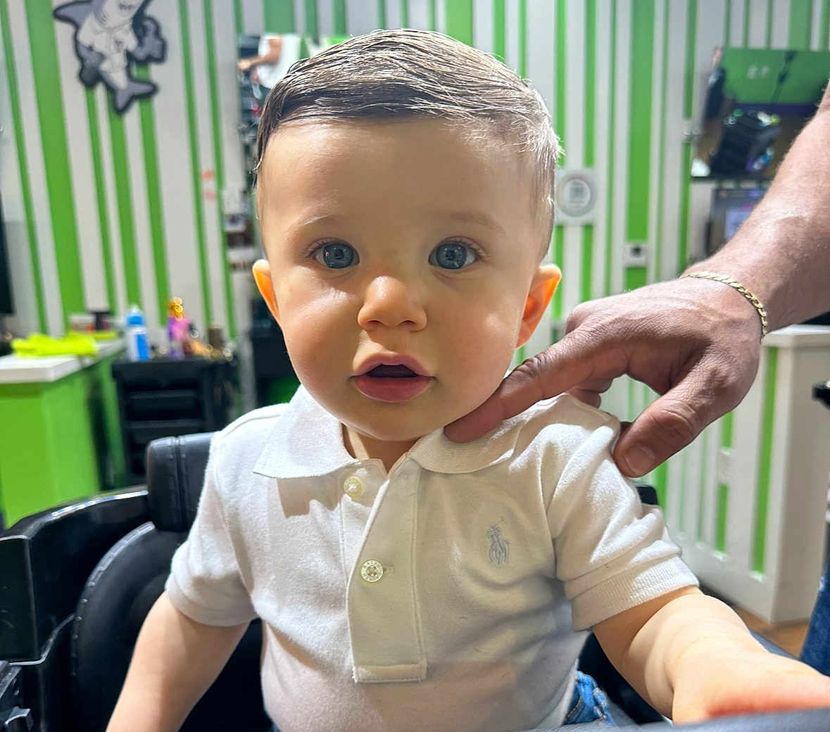 Jesus is registered to the contest to win money with this photo: toddler, child, boy, white_polo_shirt, haircut, barber_chair, adult_hand, blue_eyes, short_hair, indoor, green_wall, striped_wall, mirror, salon, person, portrait, curious, candid, clothing, furniture