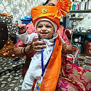 Aarvik joined the competition — help win amazing prizes! baby, child, smiling, traditional_clothing, turban, orange, white, indoor, kitchen, adult_hand, person, festive, patterned_cushion, smile, headwear, seated, pot, pan, colorful, happy