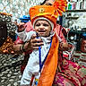 baby, child, smiling, traditional_clothing, turban, orange, white, indoor, kitchen, adult_hand, person, festive, patterned_cushion, smile, headwear, seated, pot, pan, colorful, happy