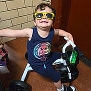Greyson joined the competition — help win amazing prizes! child, sunglasses, tricycle, indoor, toy, smile, happy, shorts, sleeveless_shirt, footwear, floor, wall, tile, play, boy, toy_vehicle, casual, person, young, fun