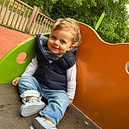 Eliot participe au concours pour gagner de l'argent avec cette photo : blond_hair, blue_eyes, casual_clothing, child, cute, daylight, fence, greenery, happy, jeans, outdoor, park, playground, shoes, sitting, slide, smile, toddler, trees, vest