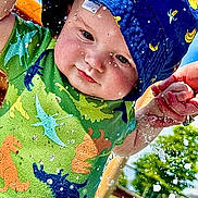 Gatlin joined the competition — help win amazing prizes! baby, child, water, splash, hat, blue_hat, dinosaur_shirt, green_shirt, outdoor, sunny, hand_holding, water_droplets, playtime, summer, happy, cute, infant, person, closeup, fun