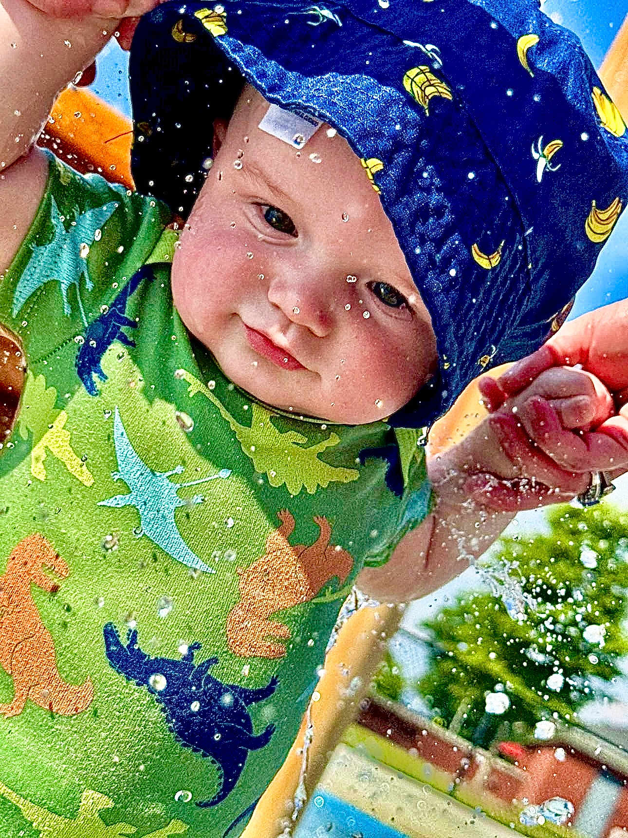 Gatlin joined the competition — help win amazing prizes! baby, child, water, splash, hat, blue_hat, dinosaur_shirt, green_shirt, outdoor, sunny, hand_holding, water_droplets, playtime, summer, happy, cute, infant, person, closeup, fun