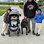 family, child, baby, stroller, dog, leash, man, boy, hoodie, toy, basketball_court, sidewalk, outdoor, trees, grass, cloudy_sky, casual_clothing, sneakers, cap, glasses