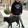 man, dog, leash, hoodie, sunglasses, sidewalk, park, slide, trees, buildings, black_dog, casual, outdoor, streetwear, pet, person, pavement, gray_pants, cap, footwear