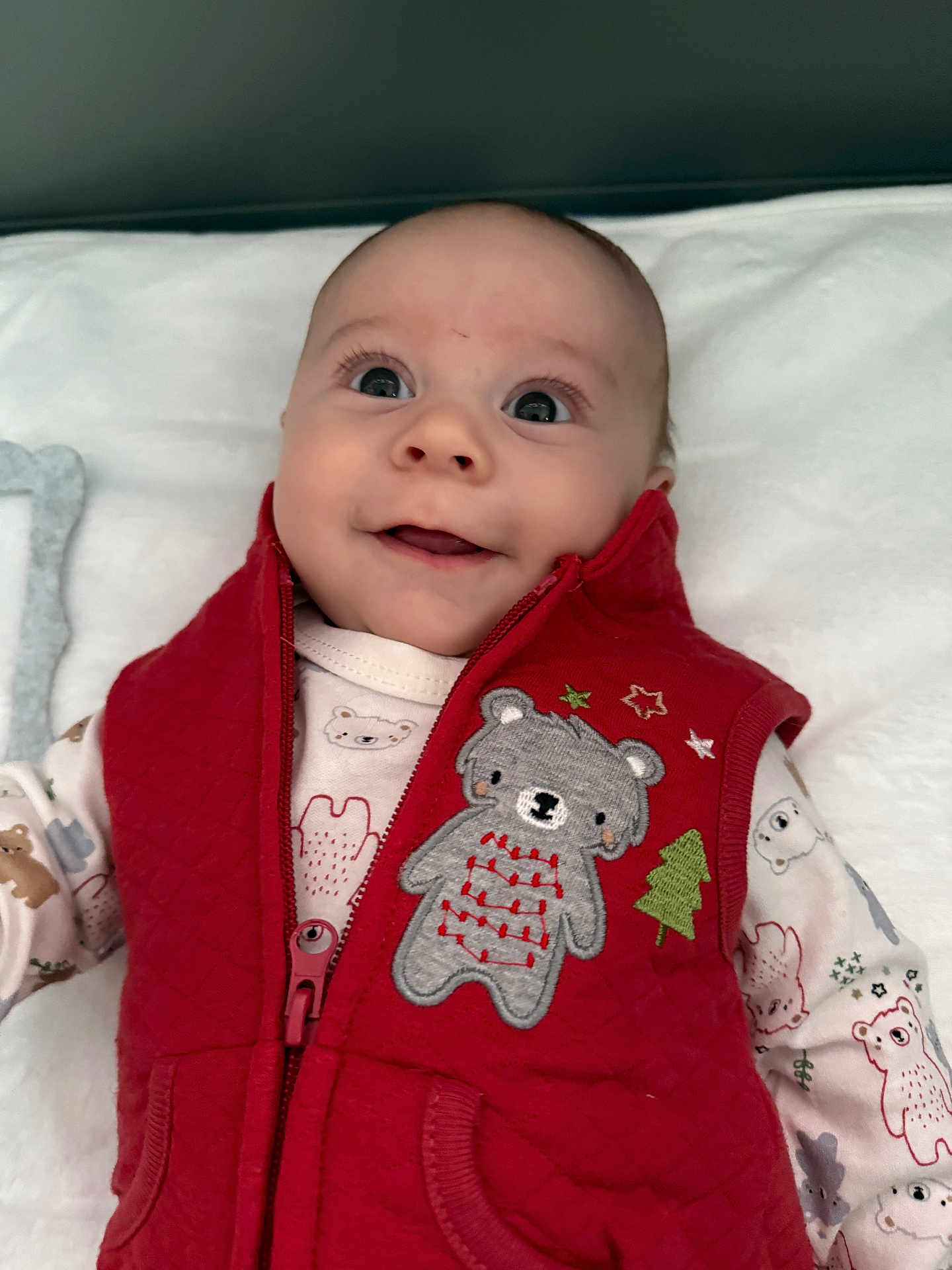 Rustyn is registered to the contest to win money with this photo: baby, infant, red_vest, bear_embroidery, smile, cute, happy, child, clothing, blanket, portrait, face, wide_eyes, indoor, soft_light, zipped_vest, patterned_onesie, young_child, close_up, cozy