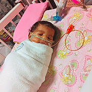 Journe'E is registered to the contest to win money with this photo: baby, infant, swaddled, blanket, crib, medical_tubes, pink_pillow, cartoon_pattern, face, resting, indoors, healthcare, child, cute, sleeping, hospital, equipment, tubing, small, peaceful