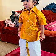 Jovanio is registered to the contest to win money with this photo: child, toddler, traditional_clothing, yellow_shirt, white_dhoti, living_room, couch, suitcase, curly_hair, bracelet, indoor, carpet, pillow, furniture, person, standing, expression, cute, home, floor