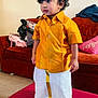 child, toddler, traditional_clothing, yellow_shirt, white_dhoti, living_room, couch, suitcase, curly_hair, bracelet, indoor, carpet, pillow, furniture, person, standing, expression, cute, home, floor