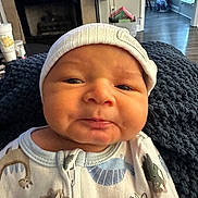 Enzo is registered to the contest to win money with this photo: baby, infant, close_up, face, hat, cap, onesie, dinosaur_pattern, blanket, indoor, cozy, expression, puckered_lips, newborn, child, person, headwear, clothing, portrait, home