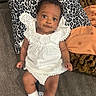 baby, infant, child, white_dress, socks, couch, pillow, patterned_pillow, leopard_print, orange_pillow, brown_pillow, carpet, indoor, person, cute, smile, face, curly_hair, relaxing, portrait