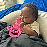baby, infant, teething_toy, pink_toy, blanket, curly_hair, indoor, window, natural_light, blue_fabric, cute, child, person, relaxing, soft_lighting, cozy, portrait, young_child, resting, baby_toy