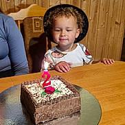 Filip is registered to the contest to win money with this photo: child, toddler, birthday_cake, candle, number_2, curly_hair, hat, wooden_wall, table, celebration, chocolate_cake, icing, sprinkles, person, indoor, happy, smile, sitting, party, young_child