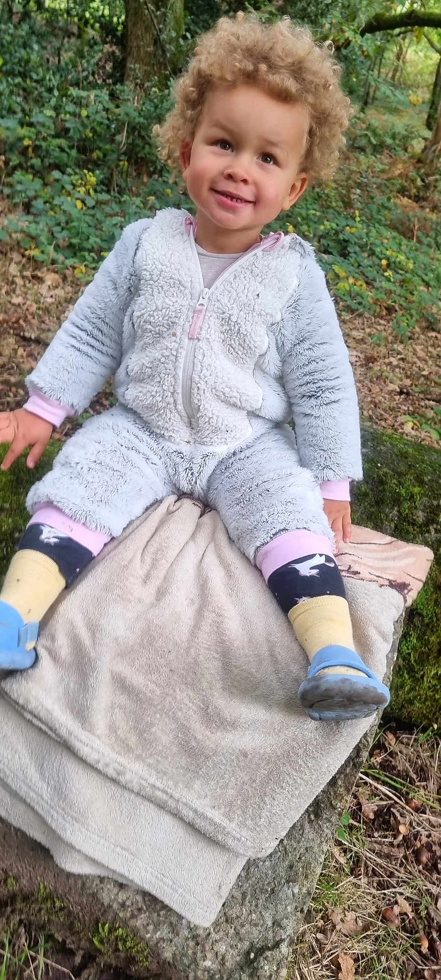 Filip joined the competition — help win amazing prizes! toddler, child, curly_hair, smiling, outdoor, forest, moss, rock, blanket, fuzzy_clothing, socks, blue_shoes, nature, greenery, happy, young_child, casual, seated, playful, daylight