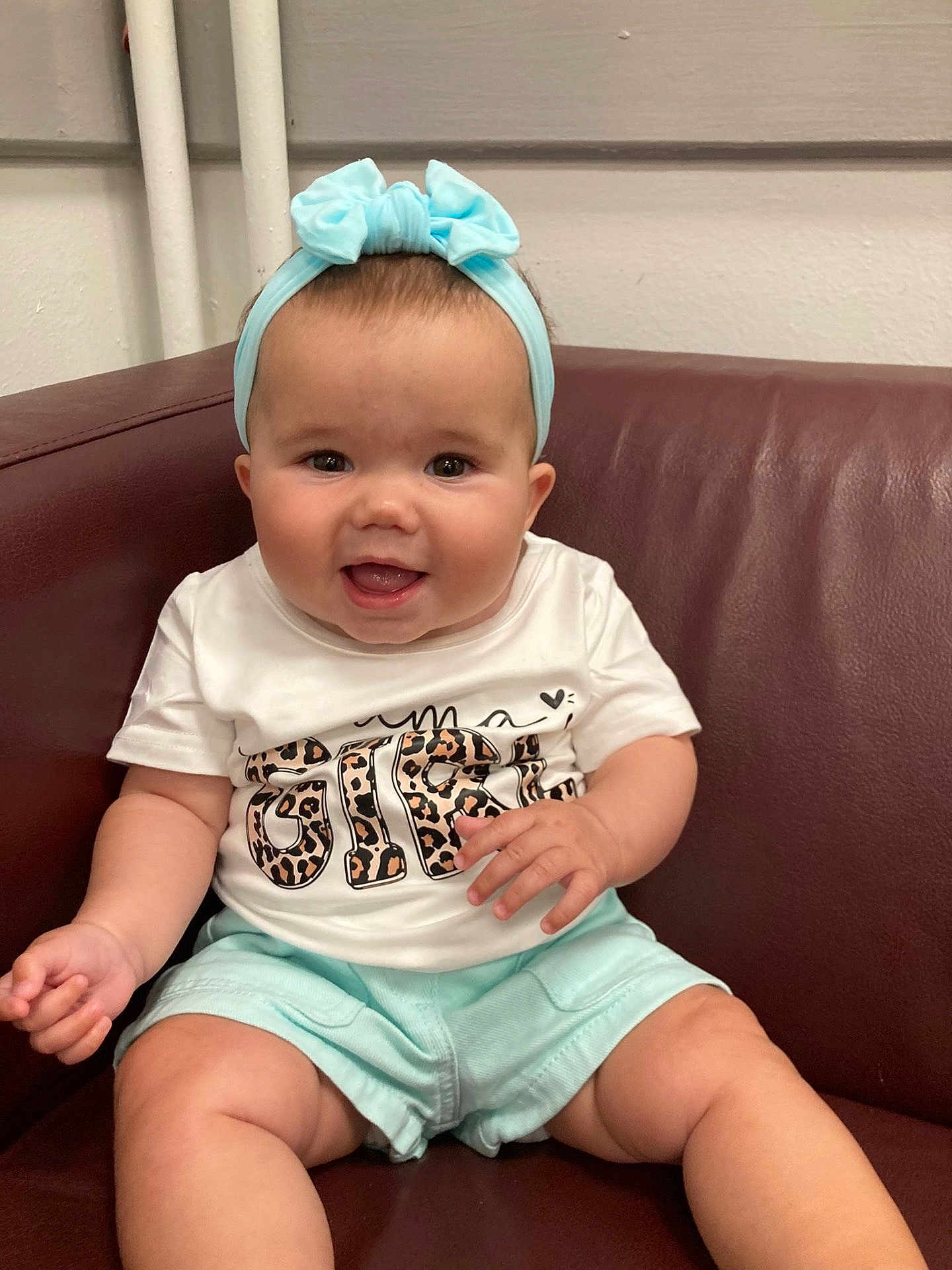 Sienna Rose is registered to the contest to win money with this photo: baby, child, smiling, headband, blue, shorts, white_shirt, leopard_print, sitting, couch, leather, happy, indoor, cute, infant, portrait, person, skin, hand, face