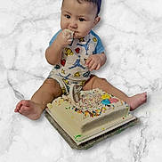 Hezekiah is registered to the contest to win money with this photo: baby, child, cake, birthday_cake, sprinkles, candle, onesie, frosting, sitting, celebration, smiling, cute, hands, feet, marble_surface, food, party, colorful, infant, eating