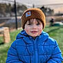 child, blue_jacket, brown_hat, outdoor, grass, winter_clothing, puffy_jacket, portrait, smiling, cold_weather, young_child, face, person, nature, daylight, cute, warm_clothing, winter, casual, innocent