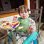 toddler, child, face, tongue_out, booster_seat, green_bib, glass_table, plate, food, spoon, onesie, tractor_pattern, chair, indoor, home, dining, window, door, baby_gate, floor
