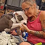 Thor is registered to the contest to win money with this photo: woman, dog, indoor, tattoos, glasses, affection, pet, casual, blanket, sitting, hand, paw, pink_tank_top, gray_dog, white_dog, cluttered_background, home, relaxed, close_up, bond