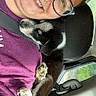 person, dog, puppy, glasses, smile, car, seat, seatbelt, window, purple_shirt, black_and_white_dog, cuddling, resting, happy, indoor, closeup, headrest, fur, paw, nature_outside
