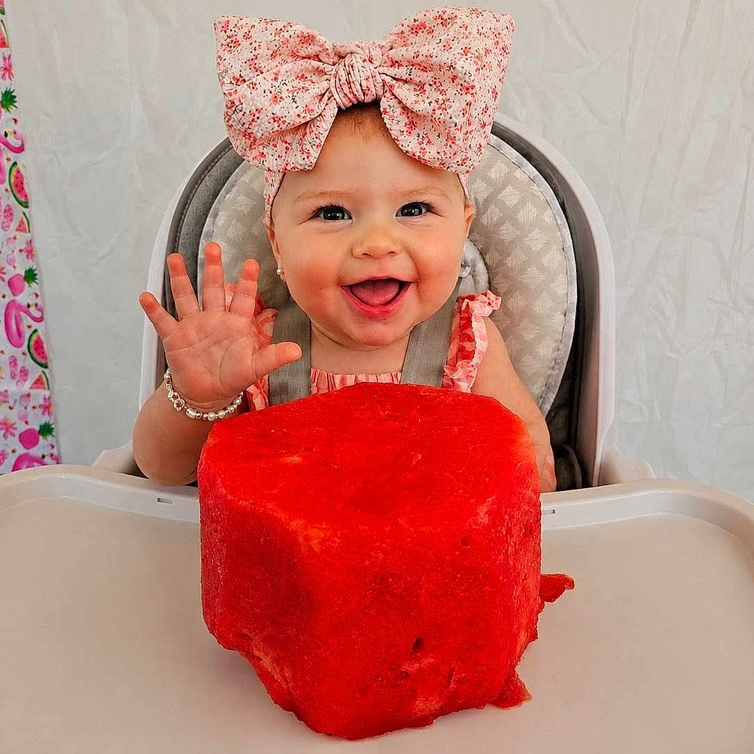 Freya is registered to the contest to win money with this photo: baby, child, high_chair, bow, watermelon, fruit, happy, smiling, hand, face, cute, food, summer, pink, patterned_curtain, jewelry, bracelet, indoor, portrait, toddler