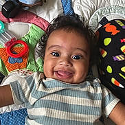 Isaiah J Anderson joined the competition — help win amazing prizes! baby, child, smiling, curly_hair, striped_shirt, lying_down, colorful_quilt, toy, blue, orange, green, black, patterned_clothing, happy, closeup, face, playful, infant, cute, indoors