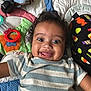 baby, child, smiling, curly_hair, striped_shirt, lying_down, colorful_quilt, toy, blue, orange, green, black, patterned_clothing, happy, closeup, face, playful, infant, cute, indoors