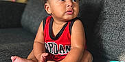 Isaiah J Anderson is registered to the contest to win money with this photo: baby, child, sitting, couch, red_clothing, jersey, indoor, curly_hair, looking_away, barefoot, skin, face, hands, foot, expression, natural_light, blurred_background, home, toy, casual