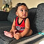 baby, child, sitting, couch, red_clothing, jersey, indoor, curly_hair, looking_away, barefoot, skin, face, hands, foot, expression, natural_light, blurred_background, home, toy, casual