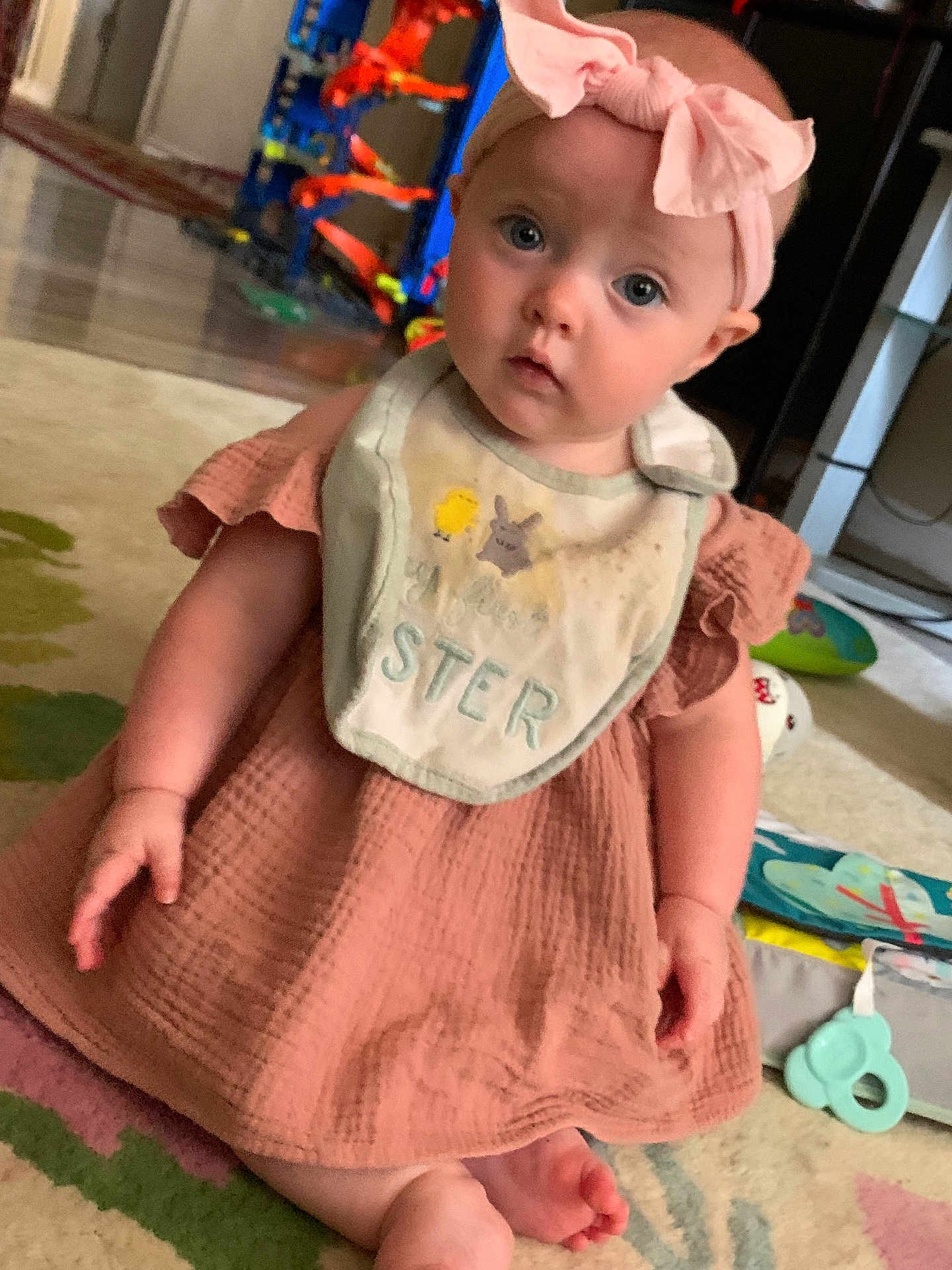 Starlite is registered to the contest to win money with this photo: baby, child, infant, pink_bow, bib, dress, carpet, toys, indoor, sitting, cute, curious, headband, footwearless, playtime, home, person, young_child, expression, floor
