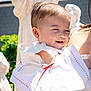 baby, child, smile, white_clothing, earring, cross_pendant, sunlight, outdoor, statue, greenery, person, portrait, cute, happy, holding, dress, infant, daylight, closeup, celebration