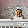 Milani is registered to the contest to win money with this photo: baby, crib, child, smiling, headband, plaid_skirt, white_clothing, pink_wall, decorative_wall, furniture, cute, portrait, indoors, infant, handrail, happy, standing, nursery, decor, baby_girl
