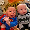 babies, batman, blanket, blue_clothing, couch, faces, family, gray_clothing, indoors, infants, phone_screen, photograph, portrait, red_cape, sitting, smiling, superhero_costumes, superman, twins, video_call