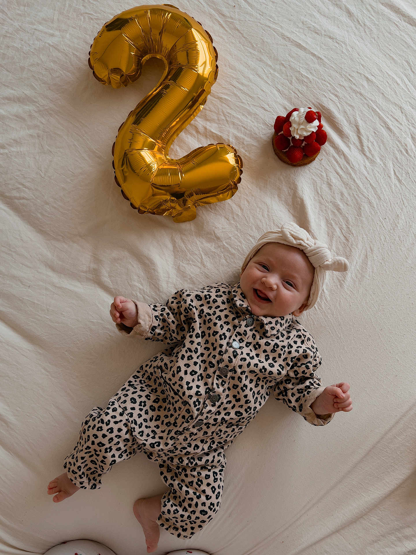 Yuna a rejoint le concours — aidez-le/la à gagner de superbes lots ! baby, infant, smiling, leopard_print, headband, balloon, number_two, cake, strawberries, white_sheet, celebration, festive, cute, happy, lying_down, clothing, portrait, indoors, child, food