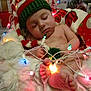 Jasper is registered to the contest to win money with this photo: baby, sleeping, christmas_lights, knitted_hat, holiday, gift_wrap, festive, cushion, bare_skin, foot, hand, indoors, decorations, red, green, white, cozy, toddler, sleep, celebration