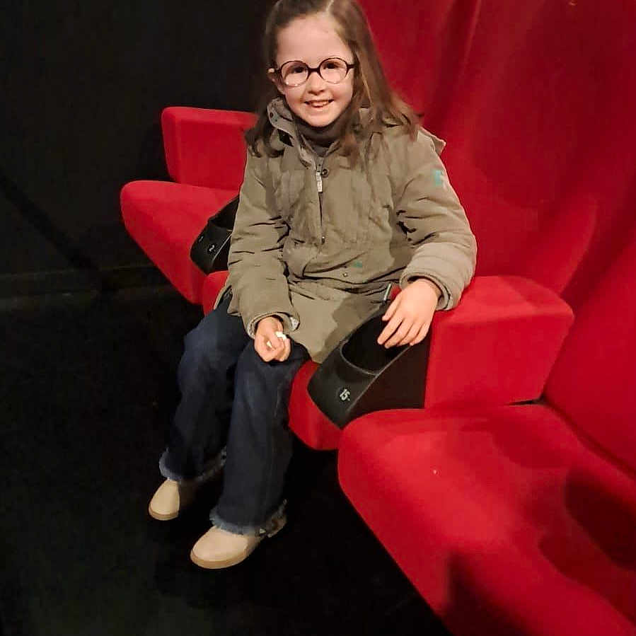Louane participe au concours pour gagner de l'argent avec cette photo : child, girl, glasses, smiling, red_seat, theater, jacket, jeans, indoor, casual_clothing, happy, portrait, seated, young, person, black_background, footwear, long_hair, leisure, cozy
