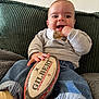 baby, blanket, child, clothing, couch, cozy, face, gilbert, grey_top, hand, happy, indoor, jeans, person, rugby_ball, sitting, smiling, texture, toy, yellow_socks