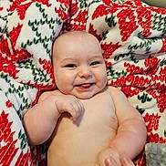 Warren is registered to the contest to win money with this photo: baby, infant, smiling, tongue_out, blanket, patterned_blanket, red, green, white, hand, fingers, skin, happy, cute, lying_down, indoors, carpet, person, cheeks, expression