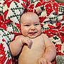 baby, infant, smiling, tongue_out, blanket, patterned_blanket, red, green, white, hand, fingers, skin, happy, cute, lying_down, indoors, carpet, person, cheeks, expression