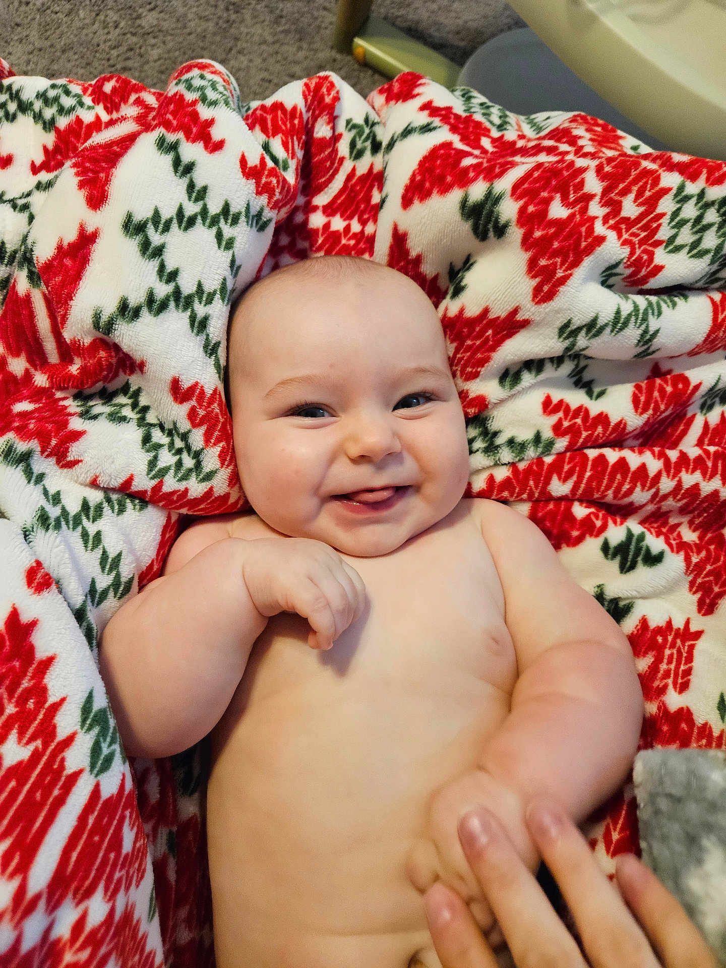 Warren is registered to the contest to win money with this photo: baby, infant, smiling, tongue_out, blanket, patterned_blanket, red, green, white, hand, fingers, skin, happy, cute, lying_down, indoors, carpet, person, cheeks, expression