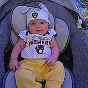 River is registered to the contest to win money with this photo: baby, infant, child, car_seat, portrait, smiling, looking_at_camera, baseball, milwaukee_brewers, sportswear, hat, onesie, yellow_clothing, purple, indoor, seated, blanket, canopy, baby_clothing, happy