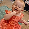 baby, child, orange_dress, smiling, finger_in_mouth, carpet, towel, indoor, room, household_items, infant, happy, cute, person, sitting, young_child, floor, face, playful, home