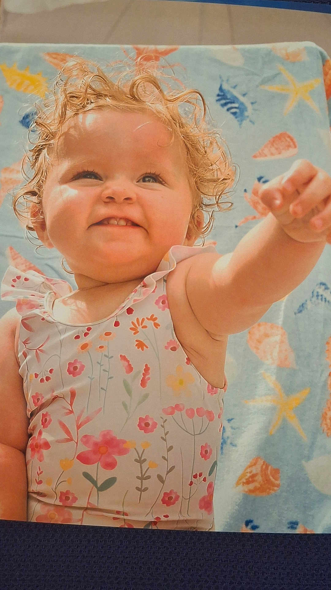 Pearl is registered to the contest to win money with this photo: toddler, child, curly_hair, smile, reaching_hand, floral_swimsuit, beach_towel, seashell_pattern, starfish_pattern, happy, outdoor, summer, cute, baby, skin, portrait, lying_down, sunlit, playful, cheerful