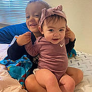 Aleah is registered to the contest to win money with this photo: baby, child, siblings, smiling, headband, onesie, bed, blanket, window_blinds, indoor, happy, cute, family, holding_hands, toddler, person, clothing, skin, face, young