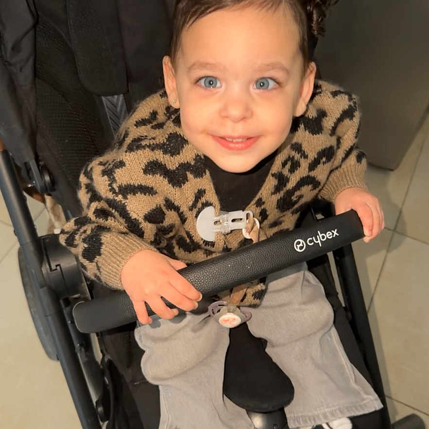 Alma participe au concours pour gagner de l'argent avec cette photo : baby_carrier, black_and_white_shoes, blue_eyes, cardigan, child, cute, cybex_brand, hair_buns, hand, indoor, jeans, leopard_sweater, pacifier, portrait, safety_bar, smiling, sneakers, stroller, tile_floor, toddler