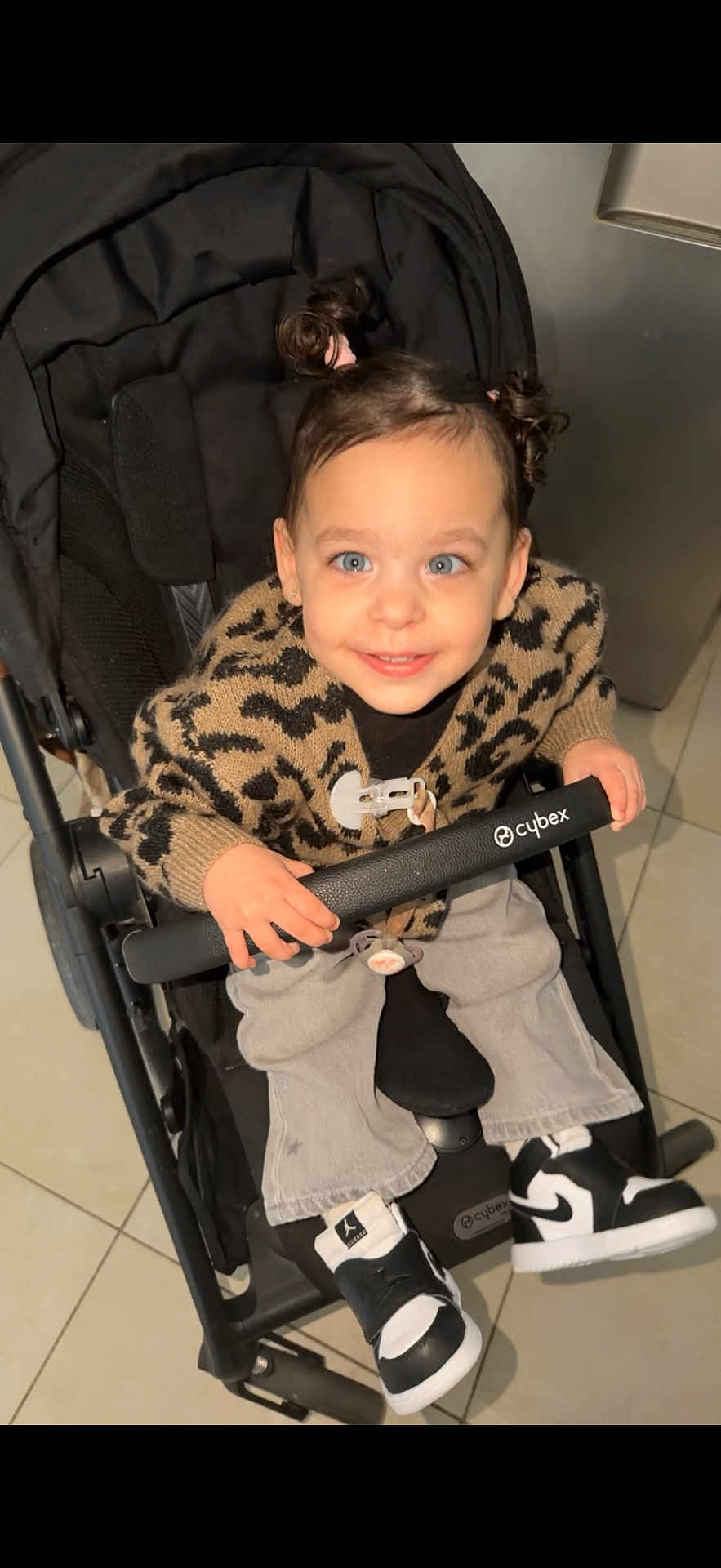 Alma participe au concours pour gagner de l'argent avec cette photo : child, toddler, stroller, smiling, blue_eyes, hair_buns, leopard_sweater, cardigan, jeans, sneakers, black_and_white_shoes, pacifier, hand, safety_bar, indoor, tile_floor, baby_carrier, cybex_brand, cute, portrait