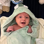 Valentin a rejoint le concours — aidez-le/la à gagner de superbes lots ! baby, smiling, hooded_towel, green_towel, plush_toy, blanket, infant, cute, happy, child, soft_texture, indoor, face, person, wrapped, cozy, newborn, cute_expression, towel, lying_down