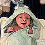 baby, smiling, hooded_towel, green_towel, plush_toy, blanket, infant, cute, happy, child, soft_texture, indoor, face, person, wrapped, cozy, newborn, cute_expression, towel, lying_down