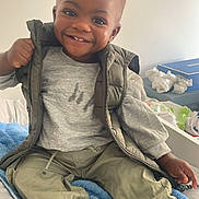 Ysaac a rejoint le concours — aidez-le/la à gagner de superbes lots ! bed, blanket, blue_blanket, casual, child, clothing, drawer, face, furniture, gray_shirt, green_vest, hair, happy, indoor, pants, person, smile, sneakers, toddler, white_blanket